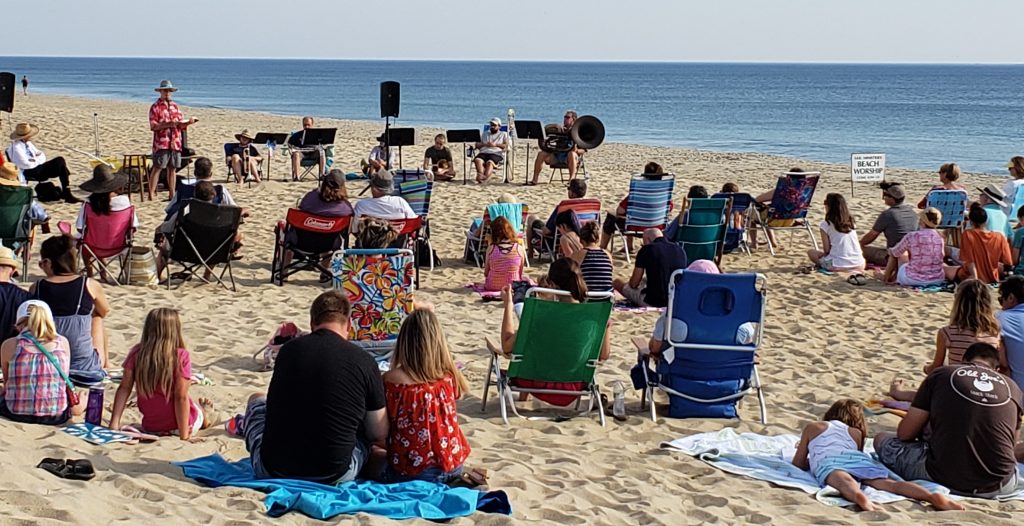 Worship service on the beach. People sitting on towels, blankets, beach chairs listening as the minister preaches the Word of God