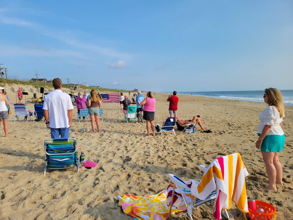 Summer morning on the Beach. Beach Worship Service. American Flag is being held for display between two people. People are standing in honor of the flag as the National Anthem is being performed by the Brass Quartet.
